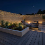 Modern outdoor patio at night with a dark wood deck, built-in planter boxes, string lights on a slatted wall, two lounge chairs, and a central fire pit table under a starry sky.