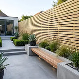 Modern outdoor patio scene with a long wooden bench set into a concrete patio, surrounded by green plants and a tall horizontal wood slat fence.