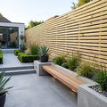 Modern outdoor patio scene with a long wooden bench set into a concrete patio, surrounded by green plants and a tall horizontal wood slat fence.