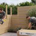 Two men installing a light wood slatted fence in a backyard setting, with one using a nail gun and the other securing a wooden panel. A branded box for 'slatted.